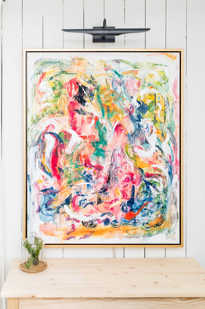 Very bright and colorful abstract art on a white vertical shiplap wall. Hangs aboive a wood table with a photo light above to highlight it. Art has gold frame called Blooming Joy from Katrina and Co. 