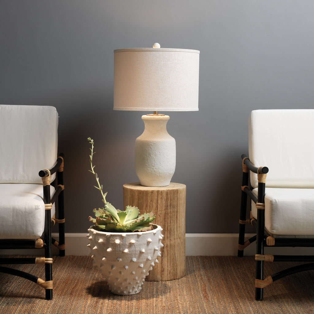 White cement table lamp with white shade on wood side table in-between two chairs against a grey wall.