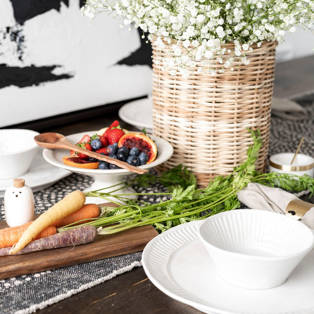 Wicker basket with flowers inside sitting on wood table with white pedestal holding fruit . White place setting with black and white table runner