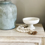 Aqua table lamp on tan nightstand in bedroom with white wall.